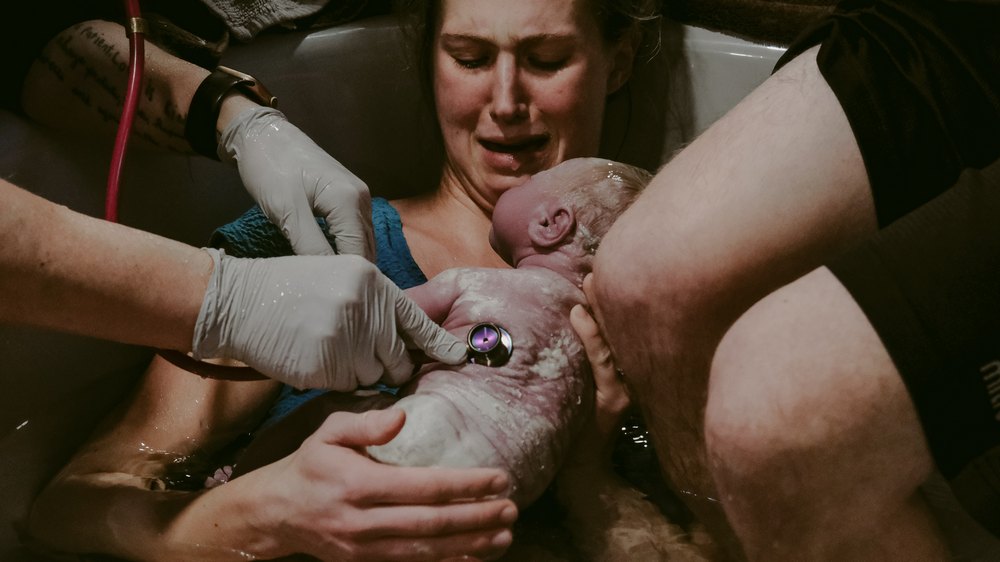 A newborn placed on its mother's chest right after birth with mother looking at it and midwives' hands checking heartbeat